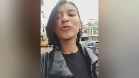 Video of a young woman with short black hair, wearing a black jacket and a septum piercing, standing in a street with buildings and parked cars in the background.