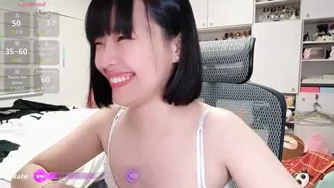 Video of a young Asian woman with shoulder-length black hair, smiling in a bedroom. She wears a white bra, with purple nipple covers. The room has a bed, a chair, and a shelf.