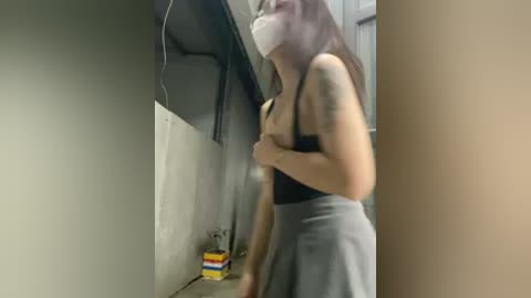 A blurry video shows a woman in a black tank top and gray skirt, wearing a face mask, standing in a dimly lit, graffiti-covered hallway with a yellow box and a black bag in the background.