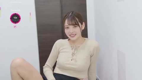 Video of an Asian woman with fair skin and straight, dark hair in a side part, wearing a beige long-sleeve top and black skirt. She sits against a white wall with a wooden door behind her.