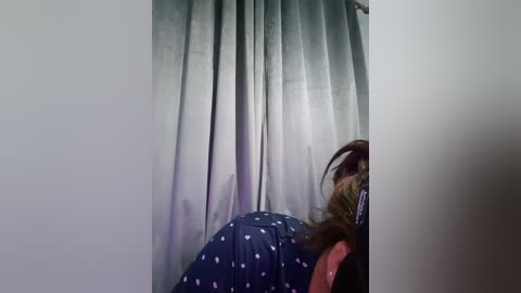 A video of a person with brown hair wearing a dark blue shower cap with white polka dots, standing in front of a light gray curtain.