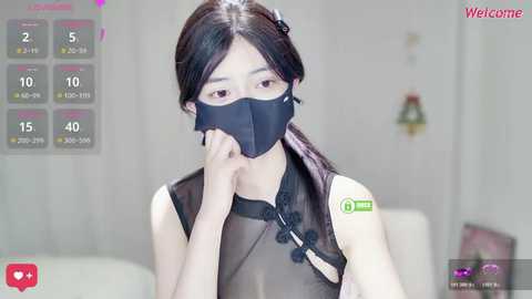 Video of an East Asian woman with long black hair, wearing a black mask, black sleeveless top, and a green and white badge, in a blurred indoor setting.