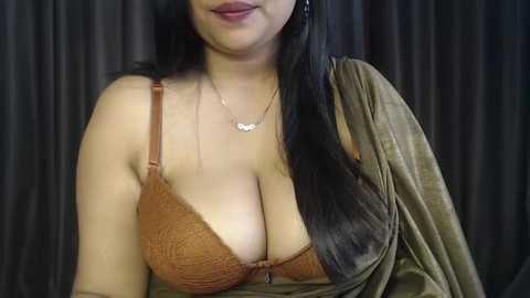 A video of a light-skinned woman with long black hair, wearing a brown lace bra that reveals ample cleavage, and a draped olive-green fabric, against dark, vertical curtains in the background.
