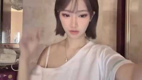 Video of a young Asian woman with short black hair and fair skin, wearing a white off-shoulder top, standing in a dimly lit, modern bathroom with beige tiles and a glass shower door.