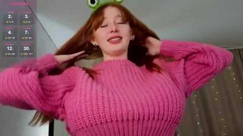 Video of a fair-skinned, red-haired woman in a pink knitted sweater, wearing a green frog hat, with a playful, wide-eyed expression, holding her hair. Background includes a calendar and curtains.