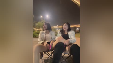 Video of two Asian women sitting on a bench at night, wearing casual clothes and holding hands. Background includes a bridge and streetlights.