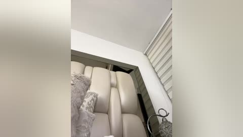 A video of a modern, minimalist bathroom with a beige, cushioned bench seat, a white sink, and a white wall with a horizontal window blind partially open.