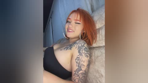 Video of a smiling, fair-skinned woman with vibrant orange hair and tattoos, wearing a black strapless top, sitting on a beige couch in a dimly lit room.
