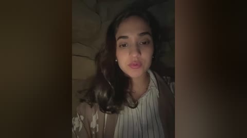 Video of a young woman with long dark hair, fair skin, and a medium build, wearing a white blouse and beige cardigan, lying on a dark stone surface.