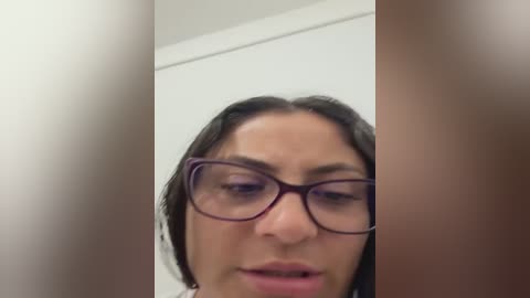 A blurry video of a woman with long dark hair, wearing red-rimmed glasses, and looking slightly upset or confused, taken from a side angle.