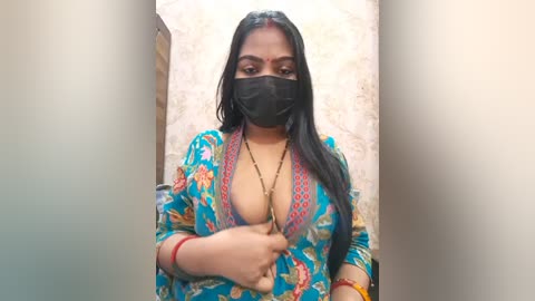 A video of a South Asian woman with long black hair, wearing a colorful floral blouse with deep neckline, black mask, and red bangles, standing against a beige textured wall.