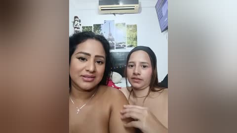 Video of two young women with light brown skin, one with long black hair, the other with long dark hair. They are topless, wearing only necklaces, in a dimly lit room with a wall of photos and an air conditioner.