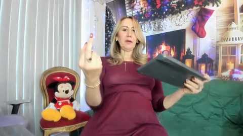 Video of a blonde woman in a maroon dress, gesturing with her right hand, holding a tablet. Background features a festive Christmas scene with a lit fireplace, garlands, and a plush Mickey Mouse toy.