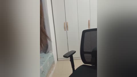 Video of a modern, minimalist office interior with a tall, slender, white wardrobe in the background, a black mesh office chair in the foreground, and a blurred, partially visible person standing to the left.