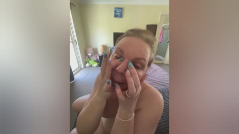 A candid video of a topless woman with fair skin, light brown hair, and blue nail polish, holding her face with both hands, taken in a bedroom with beige walls and a bed.