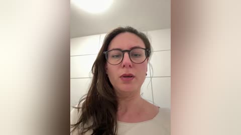 Video of a Caucasian woman with long brown hair and glasses standing in a narrow, white-tiled hallway. She wears a white top, her expression neutral.