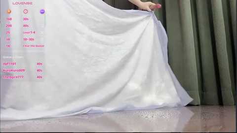 Video of a woman holding a white wedding gown with a see-through veil, standing on a polished wooden floor. Green curtains in the background.