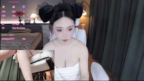 A video of a young Asian woman with fair skin and large breasts, wearing a white towel, seated in a dimly lit bedroom with a bed and a desk in the background.