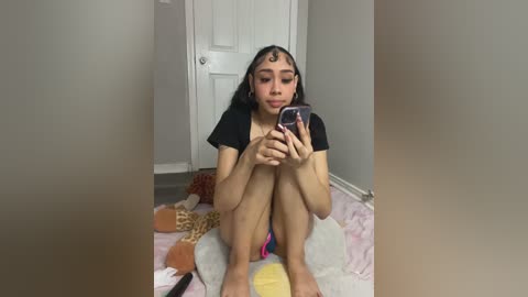Video of a young woman with long black hair, wearing a black top and shorts, sitting on a pink blanket, looking at her phone in a minimalist room with beige walls and a white door.