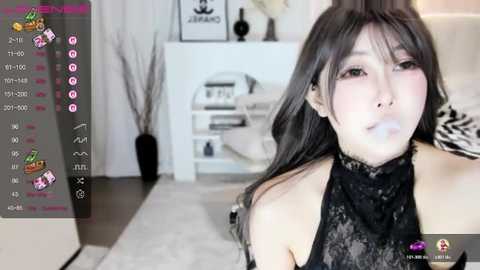 A young Asian woman with long black hair, wearing a black lace top, blowing a bubble. She's in a modern, minimalist living room with white walls, a white couch, and a white bookshelf.