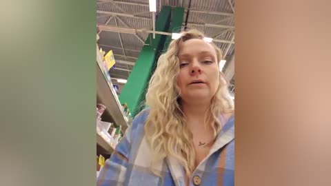 Video of a blonde woman in a blue plaid shirt, standing indoors near a green column and a metal ceiling.