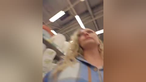 A video captures a woman with blonde hair in a blue shirt, reaching for a product in a grocery store. Shelves with various items are in the background, under fluorescent lighting. The image is slightly blurry.