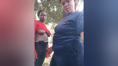 Video of two African women outdoors, one wearing a red mesh top, the other a dark T-shirt, both with large breasts. They stand in front of trees, with sunlight filtering through the leaves.