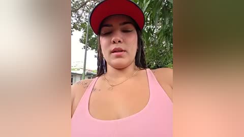 Video of a Latina woman with medium skin tone, dark hair, wearing a pink tank top and red visor, standing outdoors with green foliage and blurred residential buildings in the background.