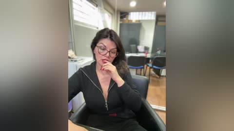 Video of a middle-aged woman with glasses and a black zip-up hoodie, seated in a modern office, pensively biting her lower lip.