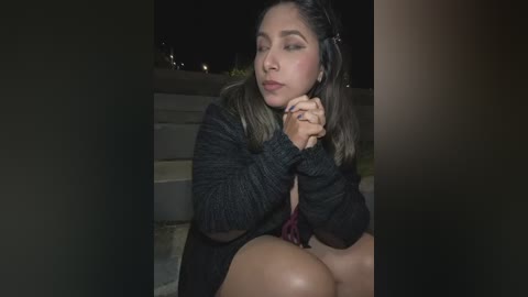Video of a Latina woman with medium skin tone and long dark hair, wearing a dark knit cardigan, sitting outdoors at night, making a pouty face, hands clasped in front.