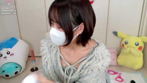 Video of an Asian woman with short brown hair, wearing a white mask, gray sweater, and pink glasses, seated at a desk with Pikachu and Eevee plushies, scissors, and a pink vibrator nearby.