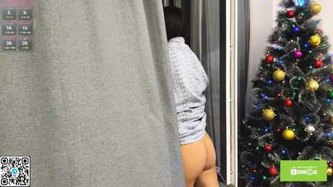 Video of a woman's buttocks partially exposed through a doorway, wearing a short, patterned dress, next to a decorated Christmas tree.