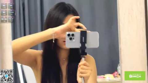 Video of a young Asian woman with long black hair, holding a smartphone with a selfie stick, partially covering her face with her hand, wearing a black sleeveless top, in a modern indoor setting.