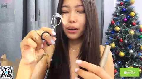 A video of a young Asian woman with long, straight black hair, wearing a sleeveless top, holding a metal fork in front of her face with a surprised expression. In the background, there's a decorated Christmas tree with colorful ornaments.