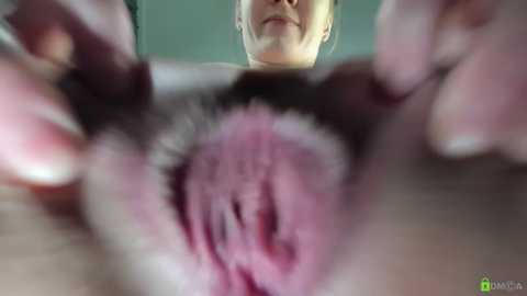 Close-up video of a woman's face and hands holding her vaginal area, highlighting pink, moist, inner labia; background blurred.