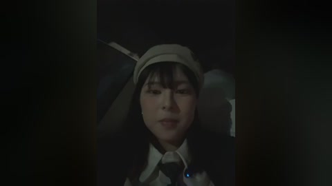 Video of a young Asian man in a dark room, wearing a light-colored hood and a white dress shirt, with a dim, shadowy background.