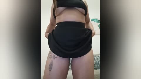 A video of a light-skinned woman with medium breasts, wearing black lingerie, lifting her skirt to reveal a thigh tattoo, standing in a room with blurred background.