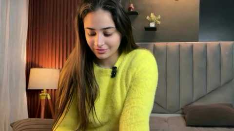 Video of a young woman with long brown hair, wearing a bright yellow sweater, sitting on a bed with a grey upholstered headboard. The background features a dark grey wall with minimalist decor.