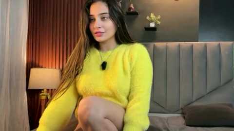 Video of a woman with long brown hair, wearing a yellow sweater, sitting on a bed with a gray headboard. Background includes a dark wall, a white lamp, and a vase with flowers.