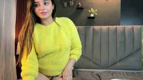 A video of a young woman with long, straight brown hair, wearing a bright yellow knit sweater, sitting on a grey tufted bench in a modern, dimly-lit room with dark walls and minimalist decor.