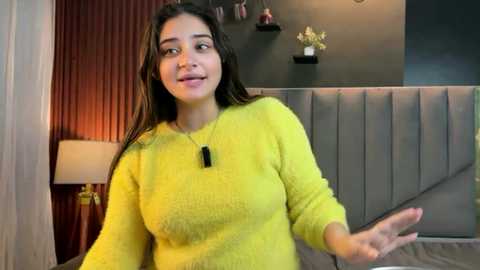 Video of a young woman with long dark hair, wearing a yellow knit sweater, standing in a modern bedroom with a gray upholstered headboard, dark walls, and potted plants.