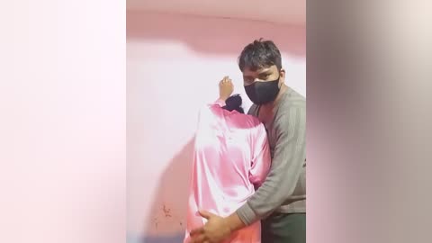 Video of a man in a gray shirt and black mask restraining a woman in a pink shirt against a pink wall in a dimly lit room.