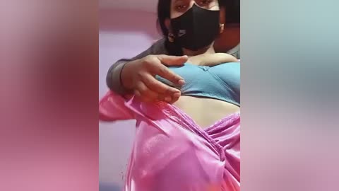 Video of a woman in a black mask, light blue bra, and pink latex dress, with a blurred background of pastel colors.