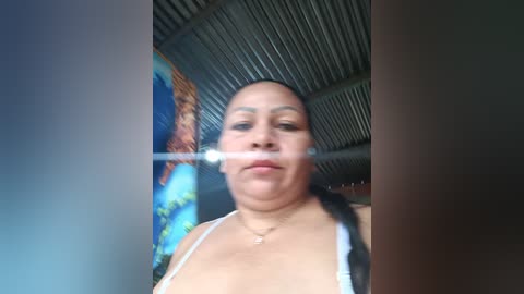 Video of a Latina woman with light brown skin, straight black hair, wearing a white tank top, standing in a room with a corrugated metal ceiling and a colorful mural on the wall.