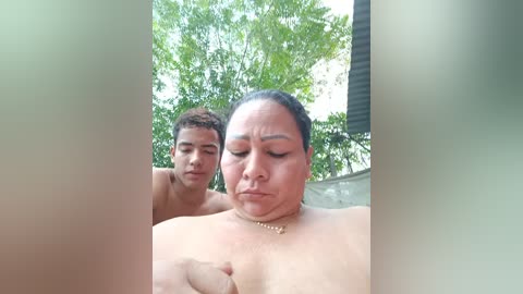 Video of a topless, middle-aged Latina woman with a pensive expression, standing next to a shirtless young Latino man with curly hair, in a tropical garden setting.