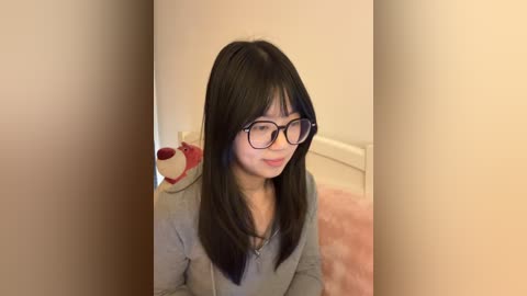Video of an Asian woman with straight black hair, wearing glasses, gray shirt, sitting on a pink carpet. She smiles slightly. Background shows beige walls and a red plush toy.
