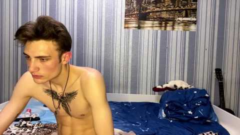 Video of a young, shirtless Caucasian male with short brown hair, wearing headphones, lying on a blue-patterned bed in a room with striped wallpaper and a framed picture of a cityscape.