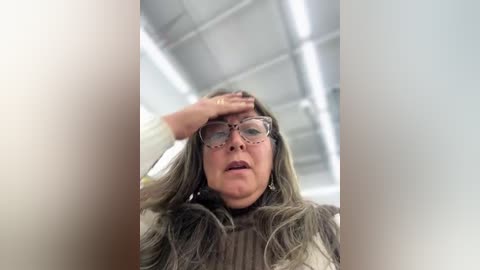 Video of a middle-aged woman with long, wavy gray hair, wearing glasses, a beige sweater, and a white shirt, touching her forehead, in an office with fluorescent lighting and a ceiling grid.