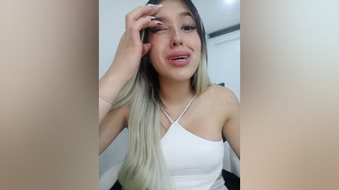 A video of a young Asian woman with long, straight, black and blonde hair, wearing a white tank top, making a playful face in a brightly lit room.