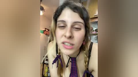 Video of a young woman with long, light brown hair, wearing a purple plaid tie and a light-colored blouse. She has a slightly flushed face and is smiling. Background shows a kitchen with cabinets and a ceiling light.